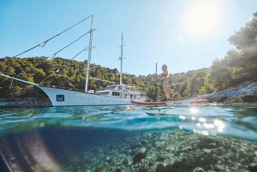 A woman stands on a paddleboard in clear blue water near a large white sailboat, with forested hills and bright sunlight in the background.