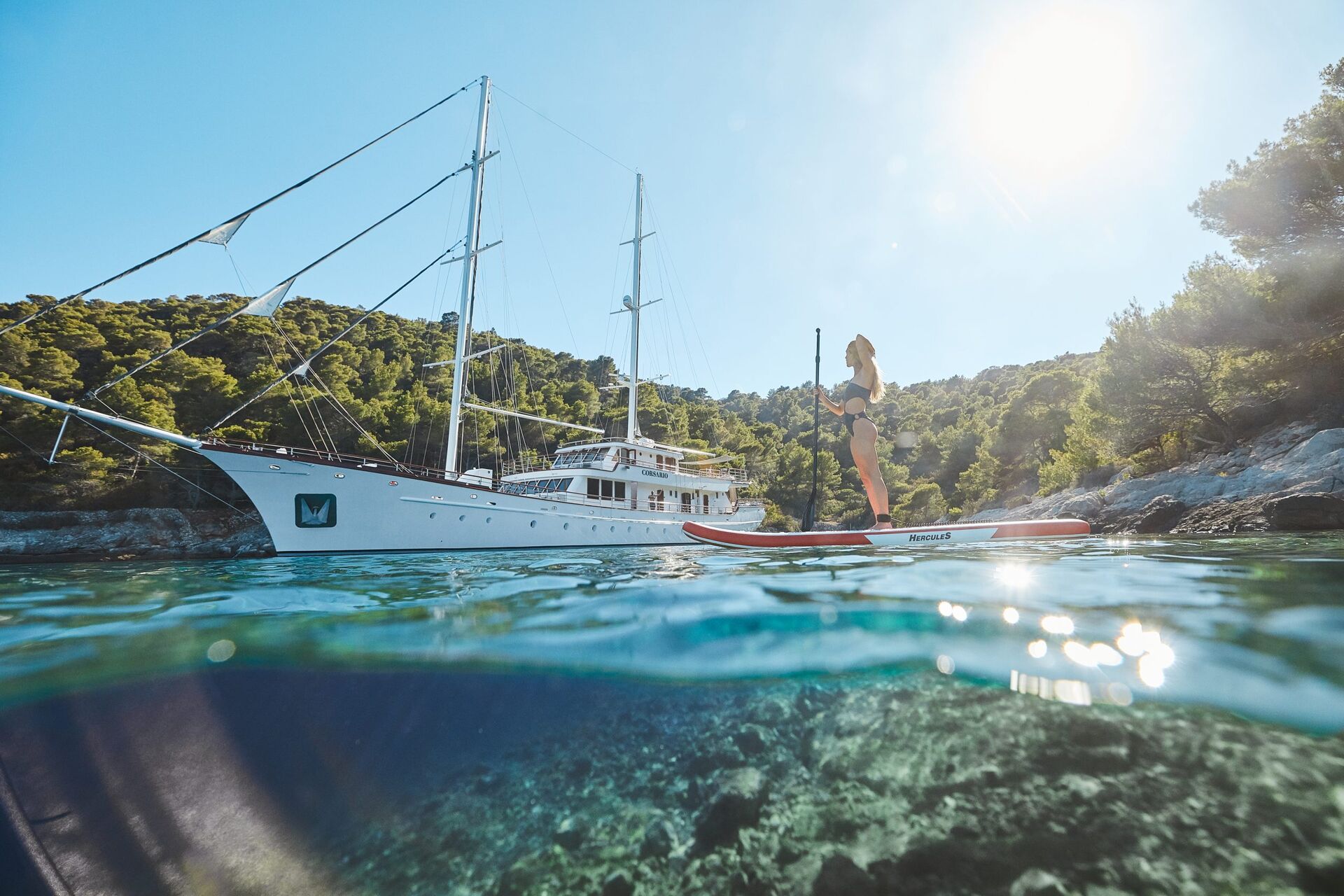 A woman stands on a paddleboard in clear blue water near a large white sailboat, with forested hills and bright sunlight in the background.
