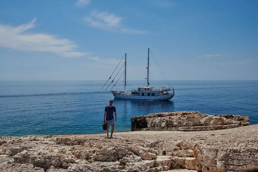 A person walks on rocky terrain by the sea, with a large sailboat anchored in the calm blue water under a partly cloudy sky.