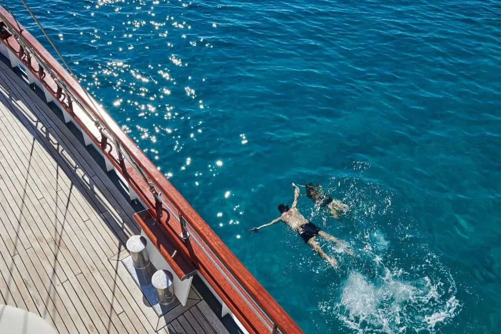 Two people swim in clear blue water next to a wooden deck of a boat, with sunlight sparkling on the seas surface.