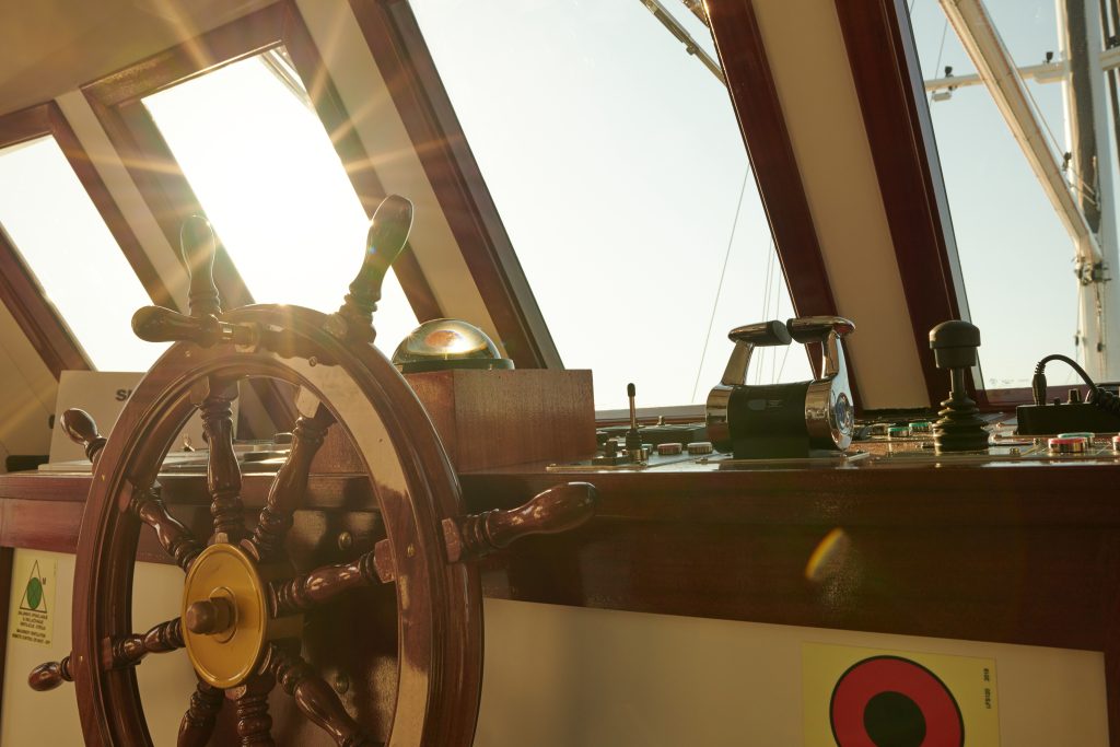 A close-up view of a ship’s wooden steering wheel and navigation controls inside the wheelhouse, with sunlight streaming through the windows and reflecting off the instruments.