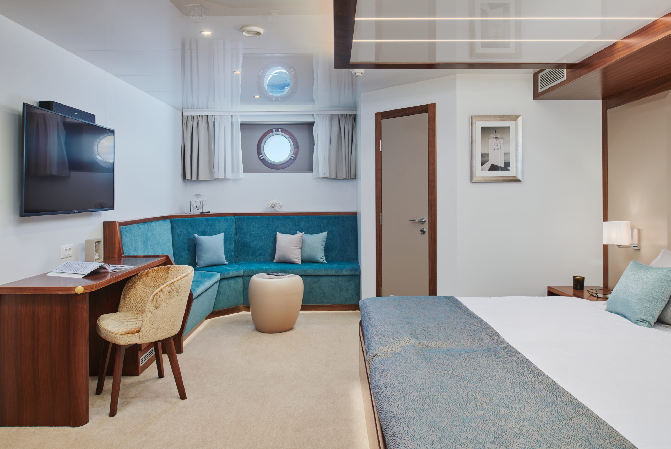 A modern cruise ship cabin features a bed, a corner sofa with teal cushions, a wall-mounted TV, a desk with a chair, a round ottoman, two porthole windows, and neutral, elegant decor.