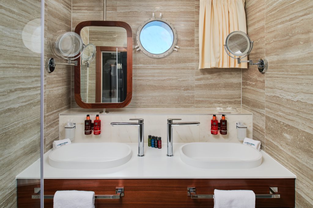 Modern bathroom with double sinks, wall-mounted faucets, two round mirrors, a porthole window, beige marble walls, and toiletries neatly arranged on the countertop. White towels hang below the sinks.