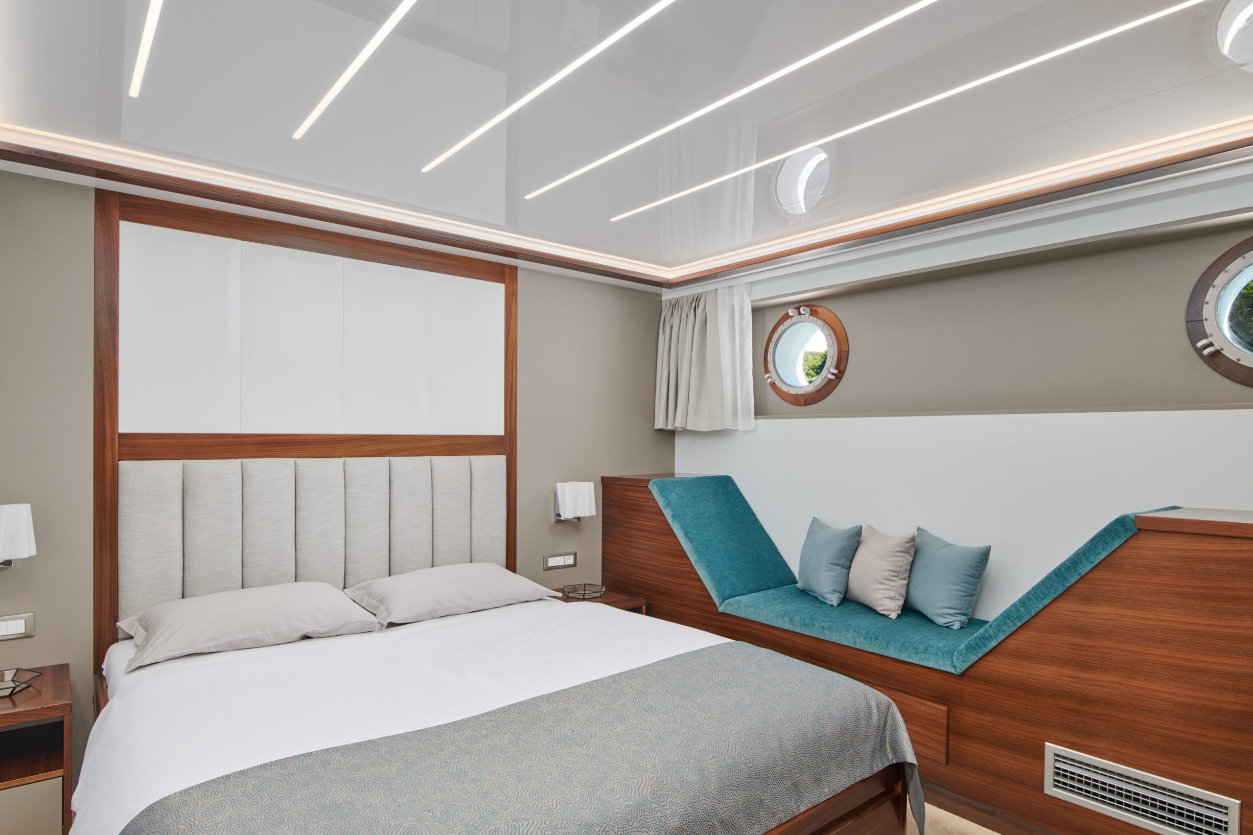 A modern bedroom with a large bed, padded headboard, small round windows, a teal cushioned bench with pillows, wood accents, and a glossy ceiling with linear lights.