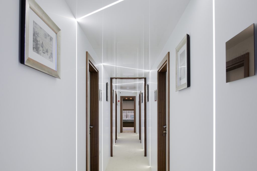 A modern, well-lit hallway with white walls, wooden doorframes, framed artwork, and bright linear ceiling lights extending the length of the corridor.