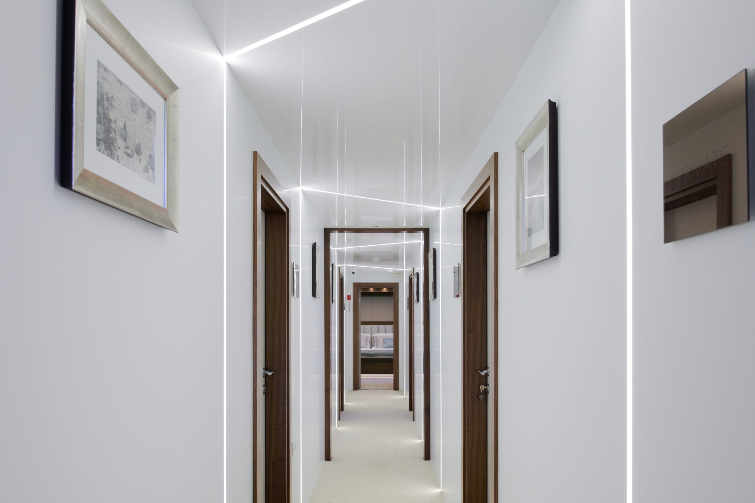 A modern, well-lit hallway with white walls, wooden doorframes, framed artwork, and bright linear ceiling lights extending the length of the corridor.