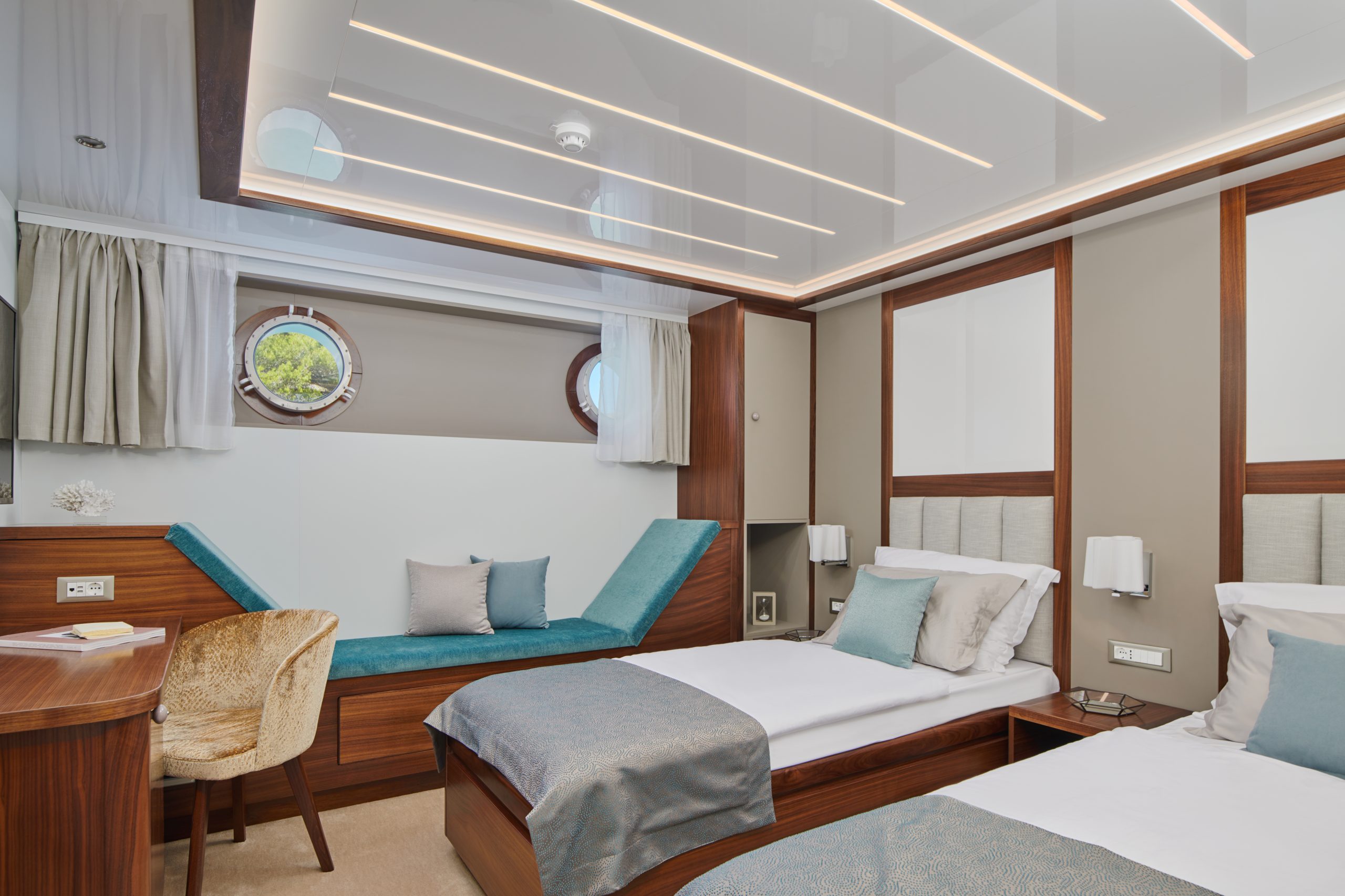 A modern cruise ship cabin with two single beds, a built-in chaise lounge by round porthole windows, a desk with a chair, and neutral-toned decor featuring wood accents and light blue bedding.