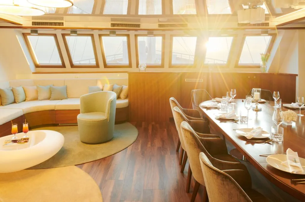 Elegant yacht interior with a sunlit dining area and a lounge featuring a round sofa, plush chair, large windows, set table with glassware, and cocktails on a white table, all under warm natural light.