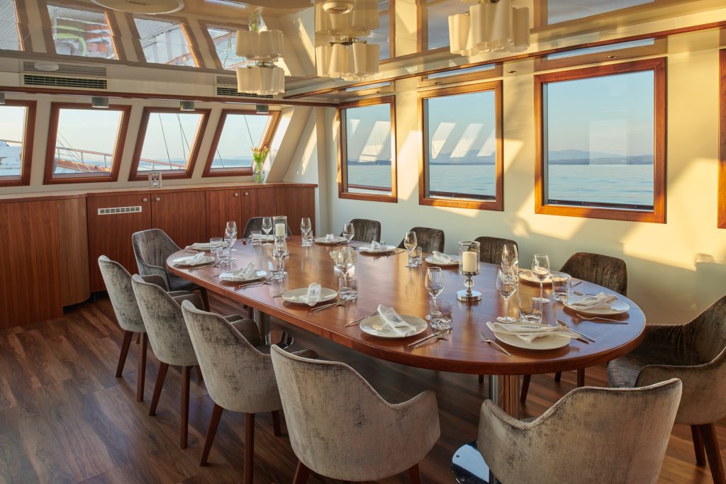 A luxurious dining room on a boat features a large wooden table set for twelve, with plush chairs, elegant tableware, and large windows offering scenic ocean views and natural light.