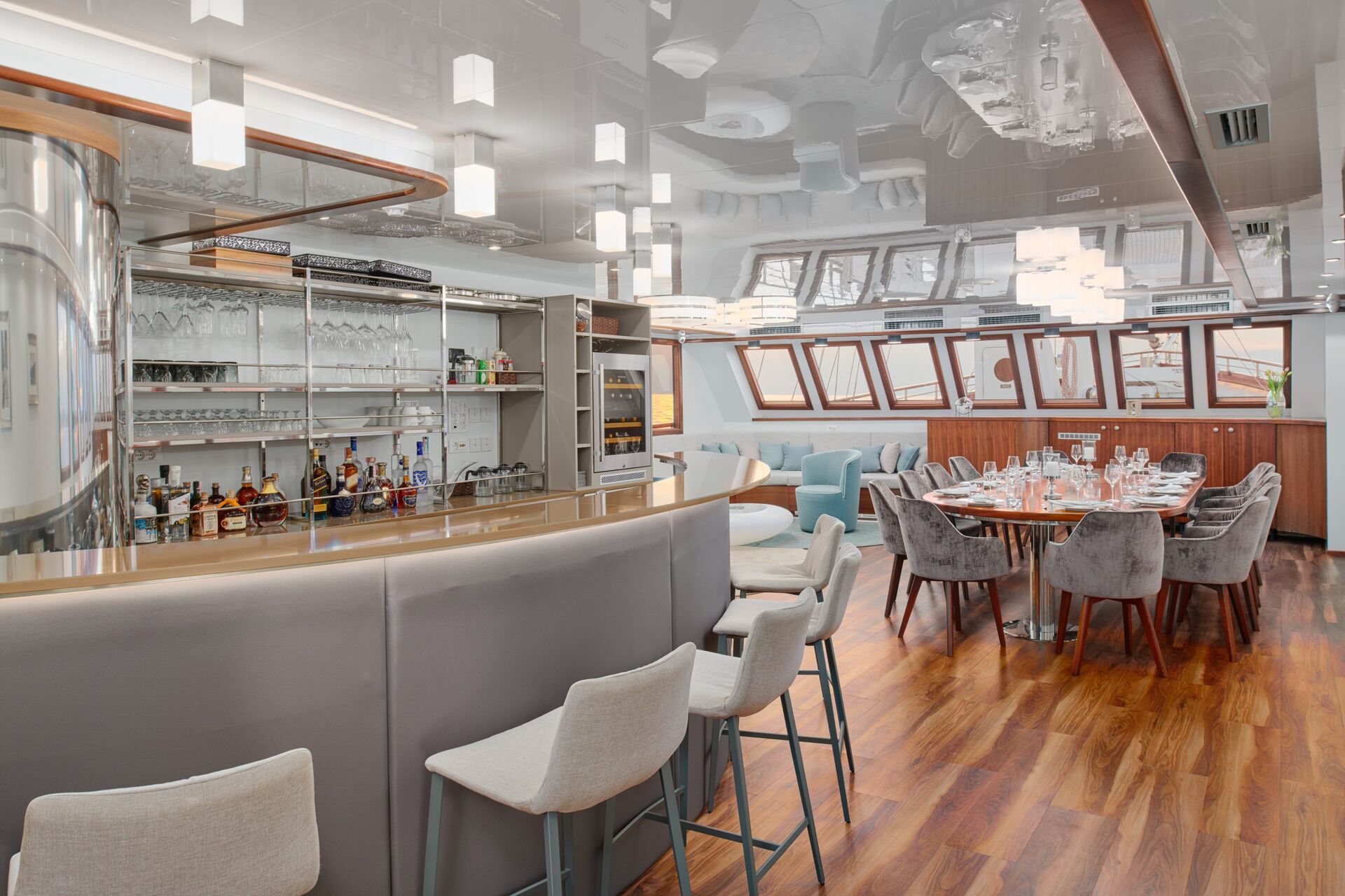 Modern yacht interior with a bar area featuring stools, shelves with glasses and bottles, and a nearby dining table set for a meal, surrounded by plush chairs and large windows letting in natural light.