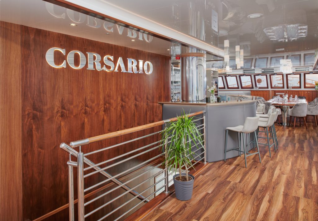 Modern restaurant interior with wood-paneled walls, a silver bar counter with high chairs, a green potted plant, and the name CORSARIO displayed in large letters on the wall. Tables are set in the background.
