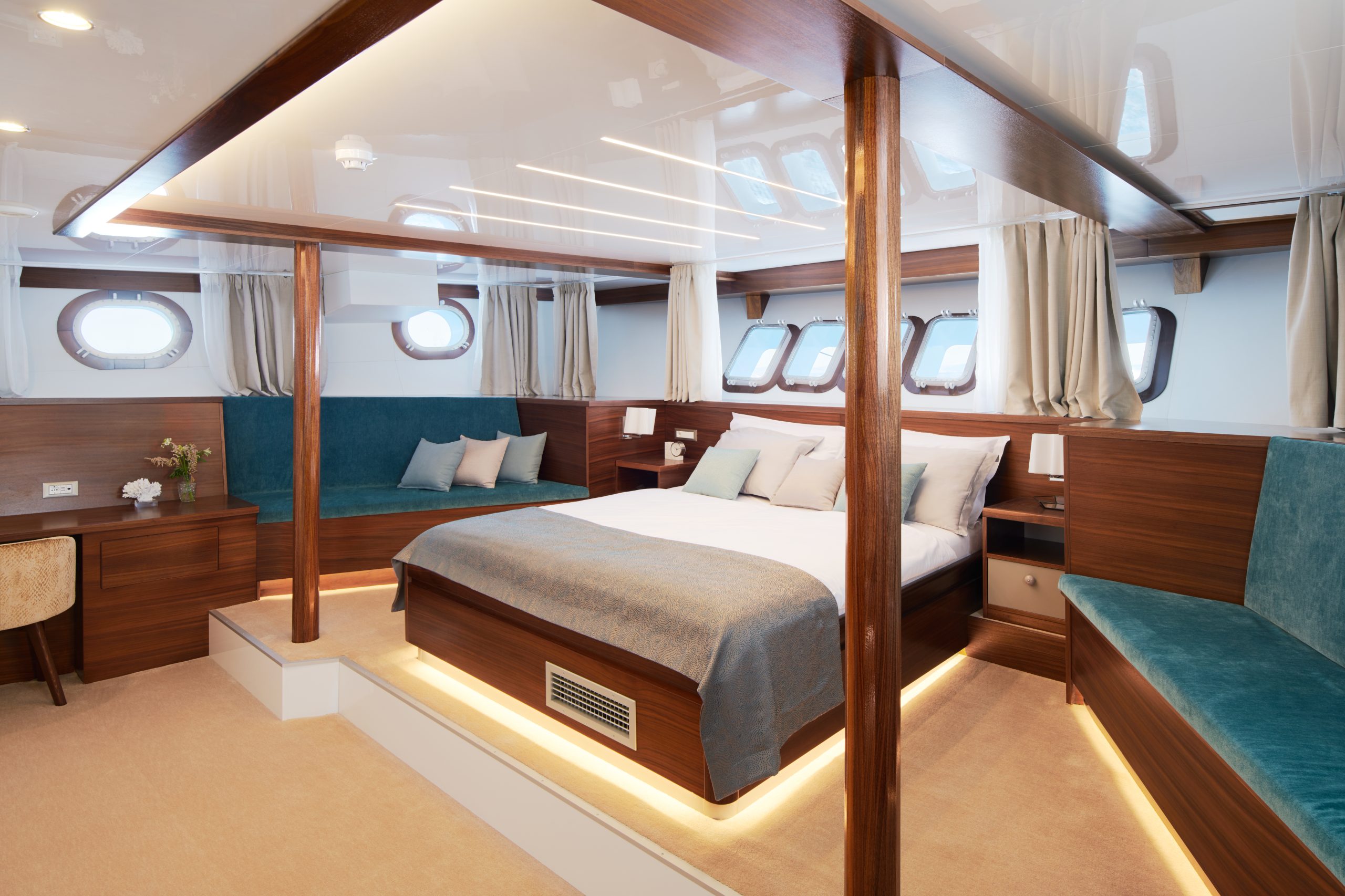 Spacious yacht cabin with a double bed, wood paneling, blue and beige decor, large windows, built-in seating, a desk with flowers, and soft lighting under the bed and benches.