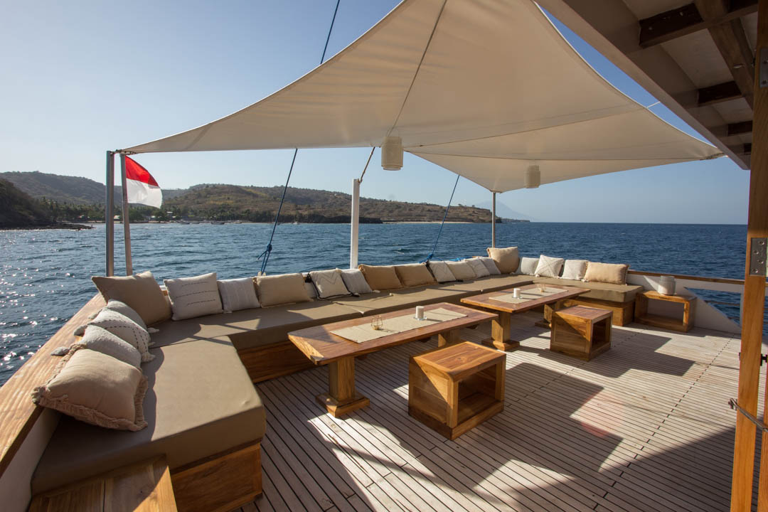 A spacious yacht deck with cushioned seating, several wooden tables, and a large white canopy, overlooking a calm sea and distant hilly coastline under sunny skies.
