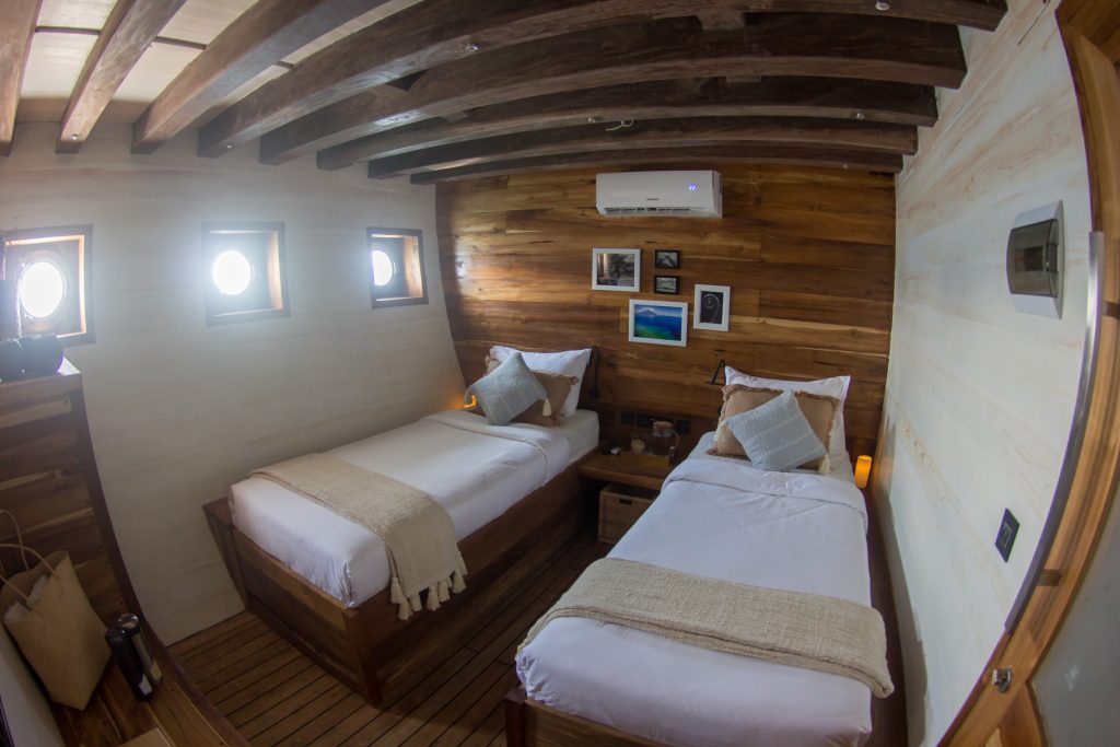A cozy cabin with two single beds, wooden walls and ceiling beams, small round windows, neutral bedding, and framed pictures above a nightstand between the beds.