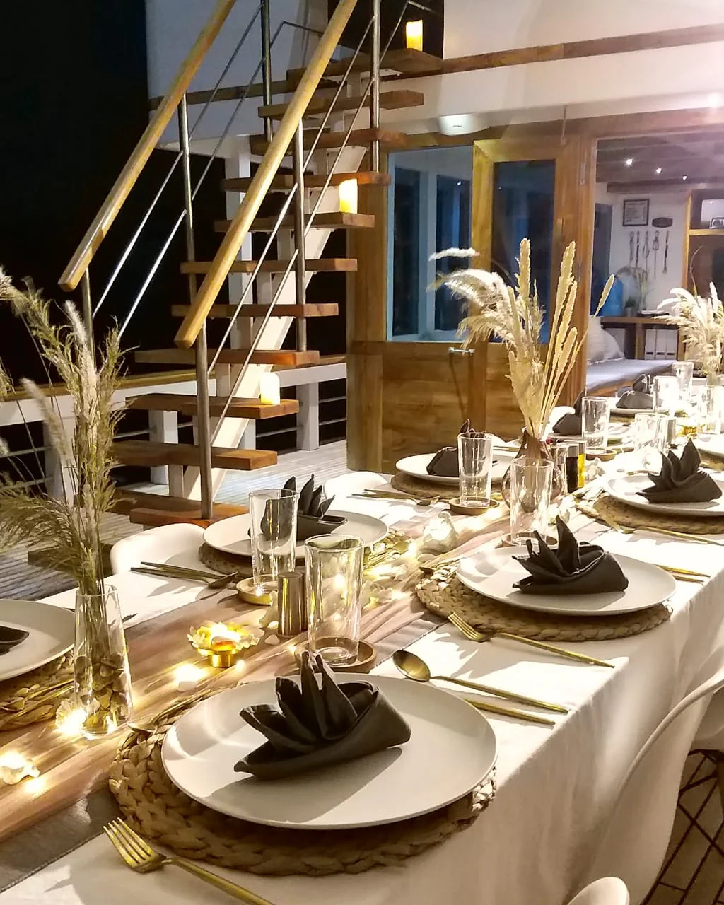 A long dining table is elegantly set with white plates, black folded napkins, gold cutlery, woven placemats, glassware, and decorative pampas grass centerpieces. Warm fairy lights run along the table. A staircase is in the background.