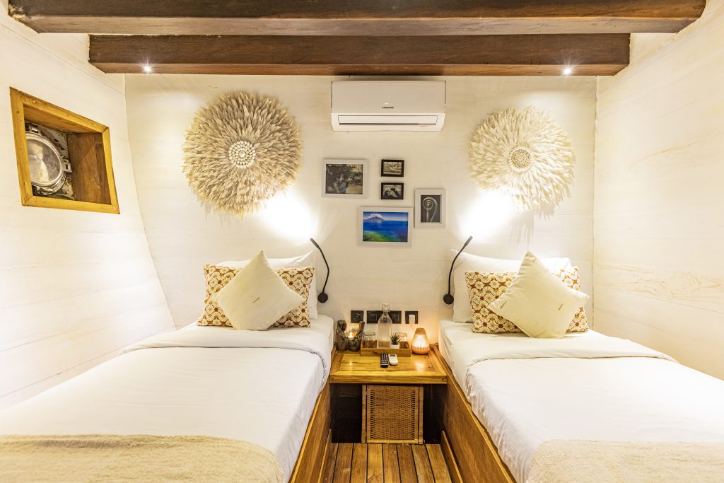 Cozy bedroom with two single beds, white bedding, decorative pillows, a wooden nightstand, wall art, feather wall hangings, and a split air conditioner above the beds. Warm lighting creates a welcoming atmosphere.