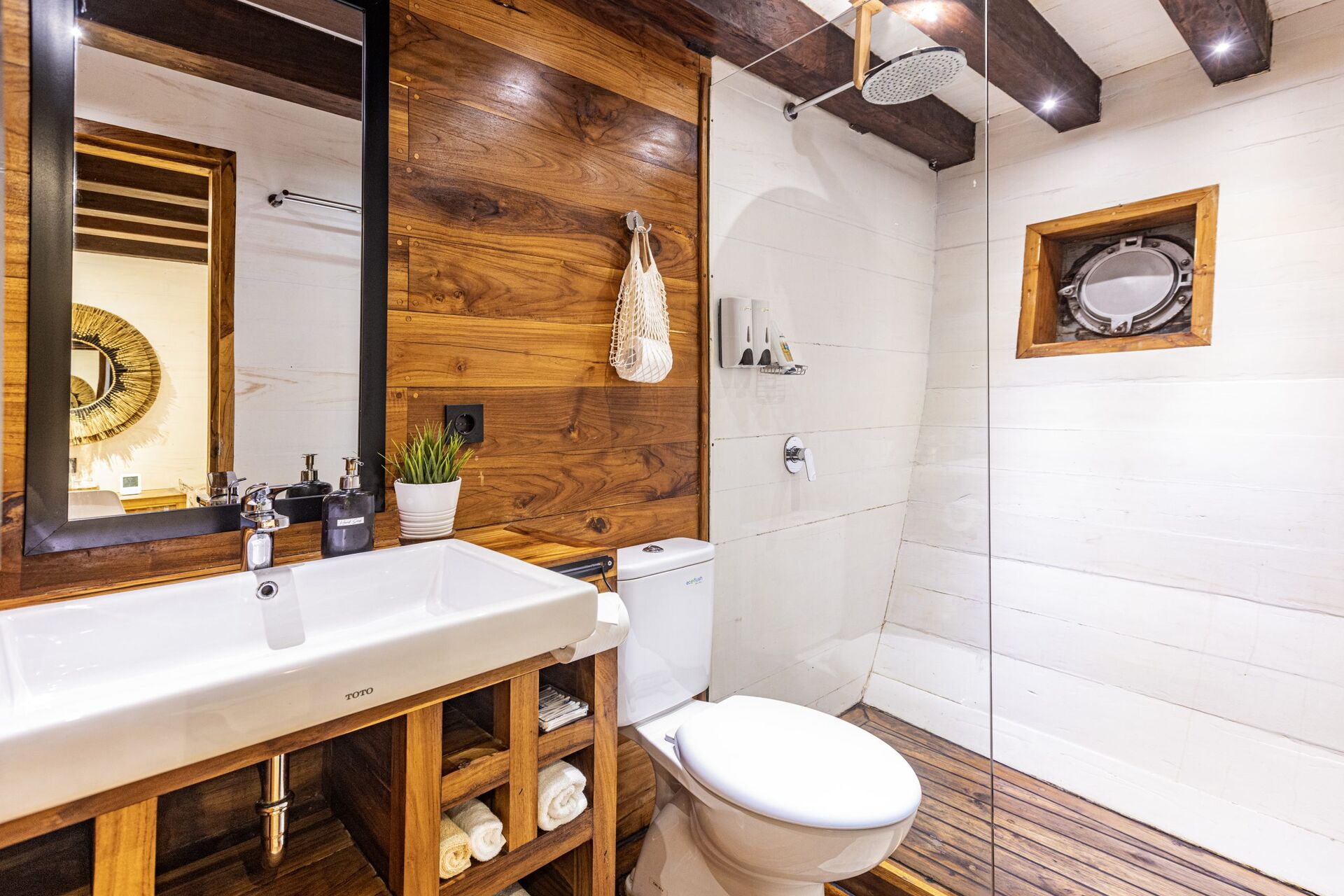 Modern bathroom with wood accents, vessel sink, mirror, white toilet, walk-in glass shower, showerhead above, small window, and towels neatly stored under the sink. A plant and toiletries decorate the sink area.