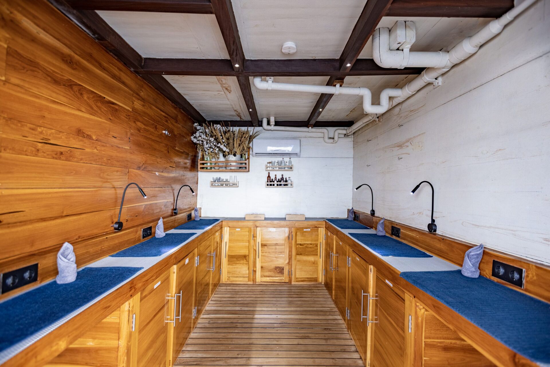 A room with wooden walls and cabinetry features eight countertop stations, each with a gooseneck faucet and rolled towel. Decorative accents and framed pictures hang on the back wall, beneath exposed ceiling pipes.
