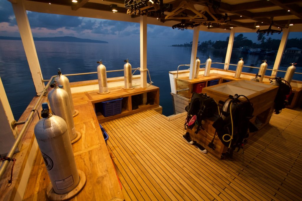 A wooden dive boat deck at dusk, equipped with multiple scuba tanks, gear benches, and clear railings, floating on calm water with a distant shoreline and cloudy sky in the background.