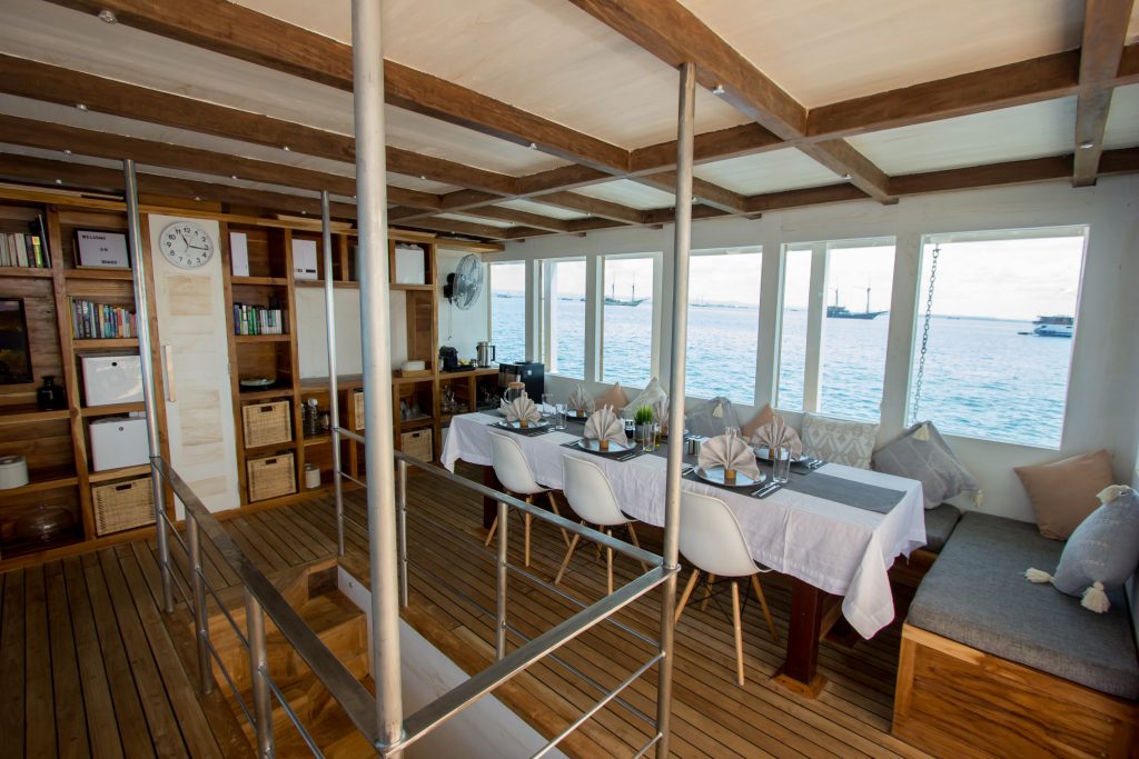 A cozy, sunlit boat interior with wooden floors and ceiling, a dining table set for a meal, bookshelves, storage baskets, and large windows showing a view of the sea and anchored boats outside.