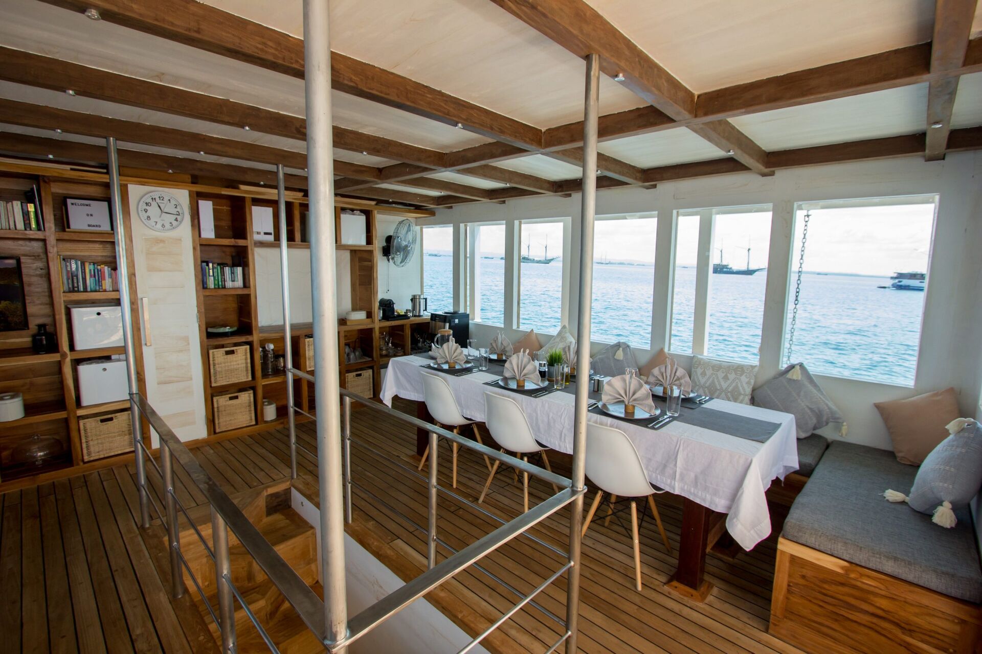 A cozy, sunlit boat interior with wooden floors and ceiling, a dining table set for a meal, bookshelves, storage baskets, and large windows showing a view of the sea and anchored boats outside.