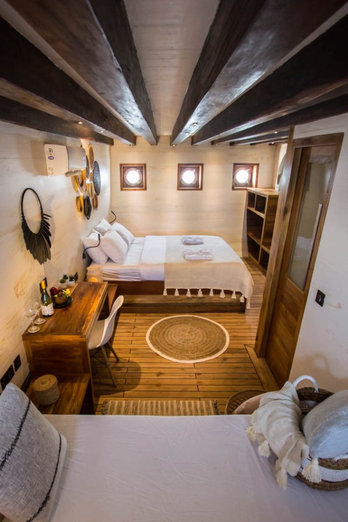 Cozy cabin bedroom with wooden beams, a double bed, a single bed, a desk with a chair, round mirrors on the wall, porthole windows, woven rug, and warm wood accents throughout.