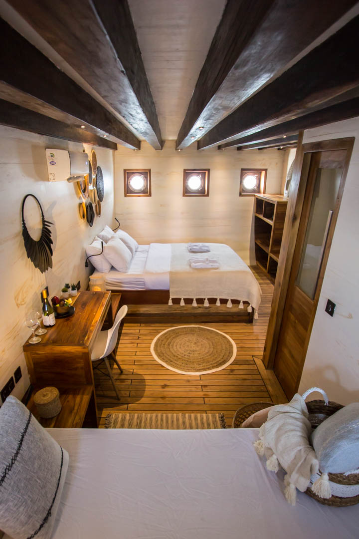 Cozy cabin bedroom with wooden beams, a double bed, a single bed, a desk with a chair, round mirrors on the wall, porthole windows, woven rug, and warm wood accents throughout.