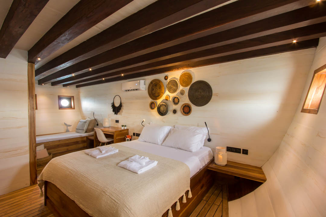 A cozy bedroom with a wooden ceiling and light-colored walls, featuring a double bed with white bedding, a desk with a chair, wall-mounted baskets for decor, and soft natural lighting.