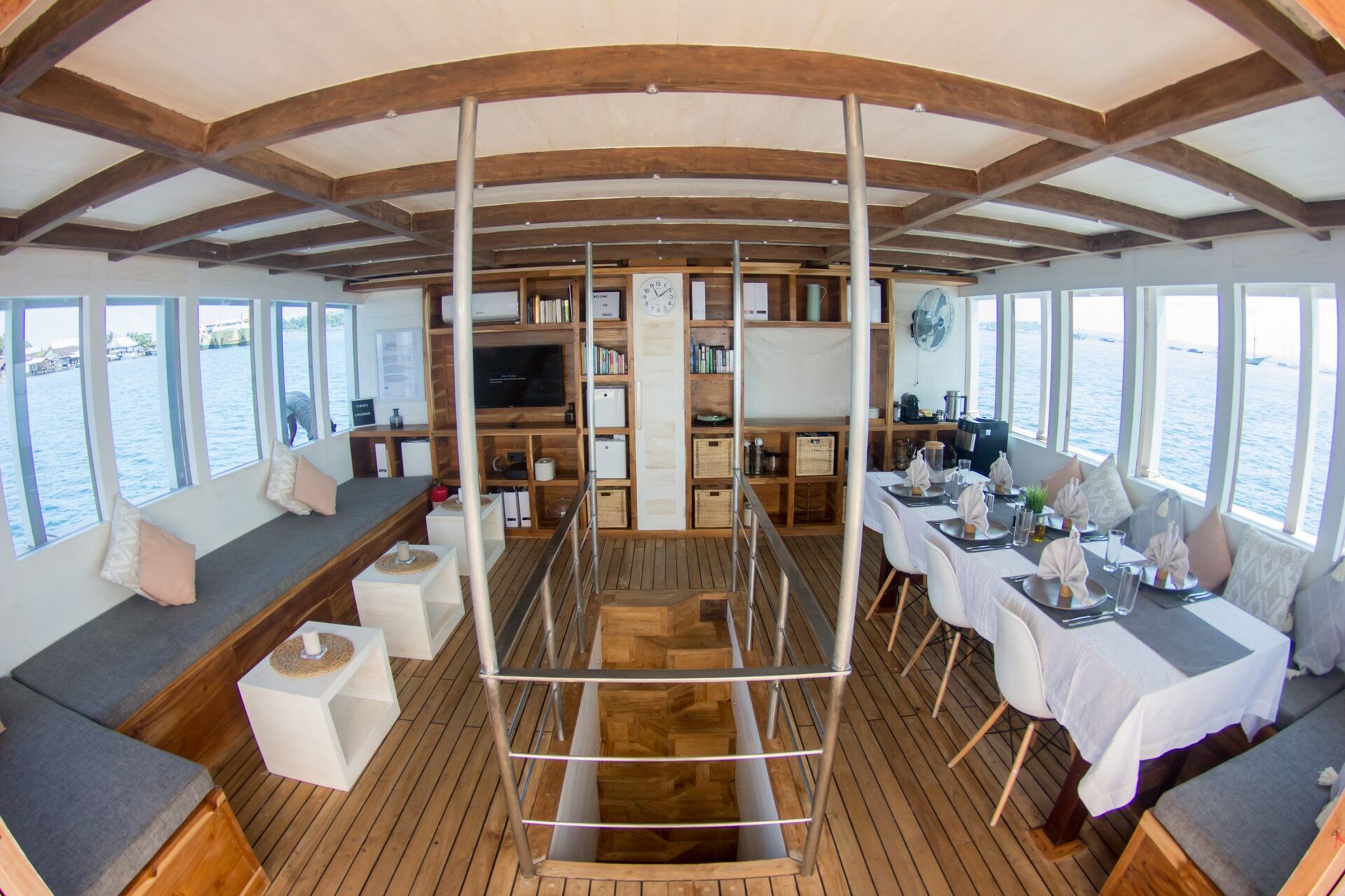 Interior of a boat with wooden floors and ceiling beams, large windows on both sides, cushioned benches, dining tables set with plates and glasses, and a staircase in the center leading below deck.