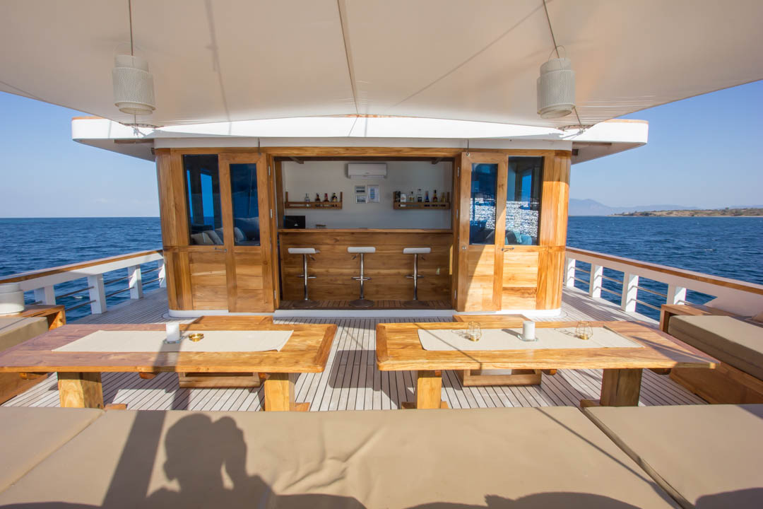 Outdoor seating area on a wooden boat deck with cushioned benches and tables facing a bar, all under a large canopy. The boat is on calm blue water with land visible in the distance.