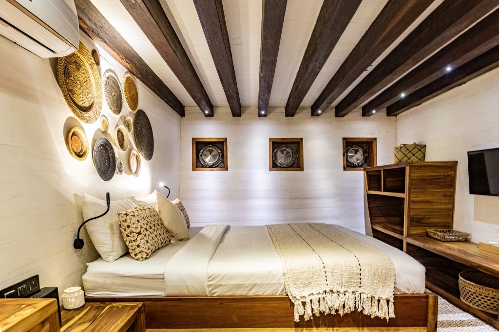 Cozy bedroom with a wooden bed, neutral bedding, woven wall art, exposed ceiling beams, four square windows, wooden shelves, and a wall-mounted TV. Ambient lighting creates a warm, inviting atmosphere.