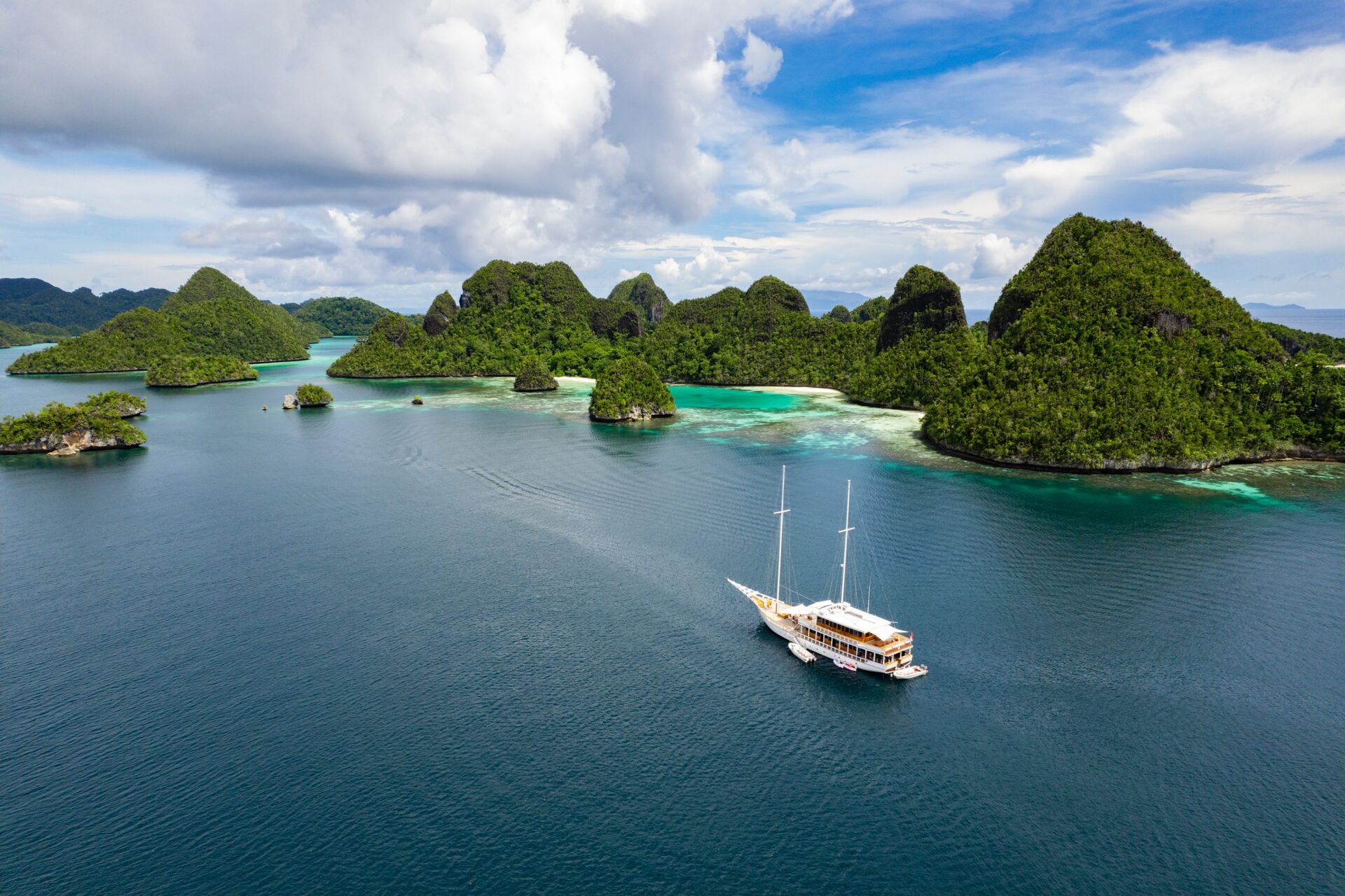 A white sailing yacht cruises through clear blue water, surrounded by lush green, rocky islands under a partly cloudy sky in a tropical seascape.