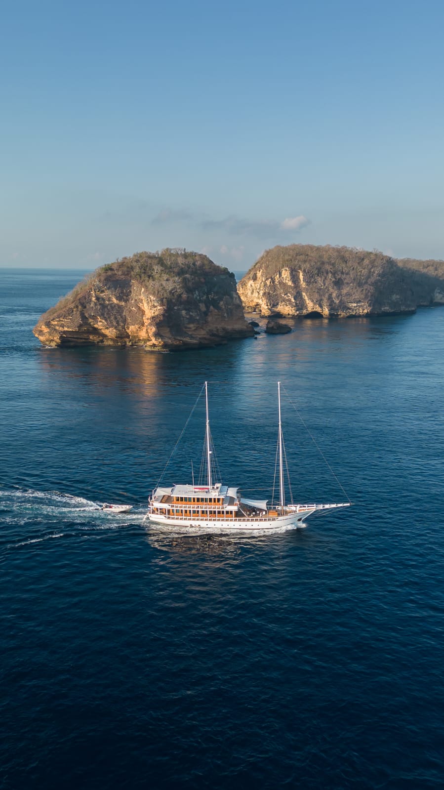 A white sailboat cruises on calm blue water near two rocky, tree-covered islands under a clear sky.