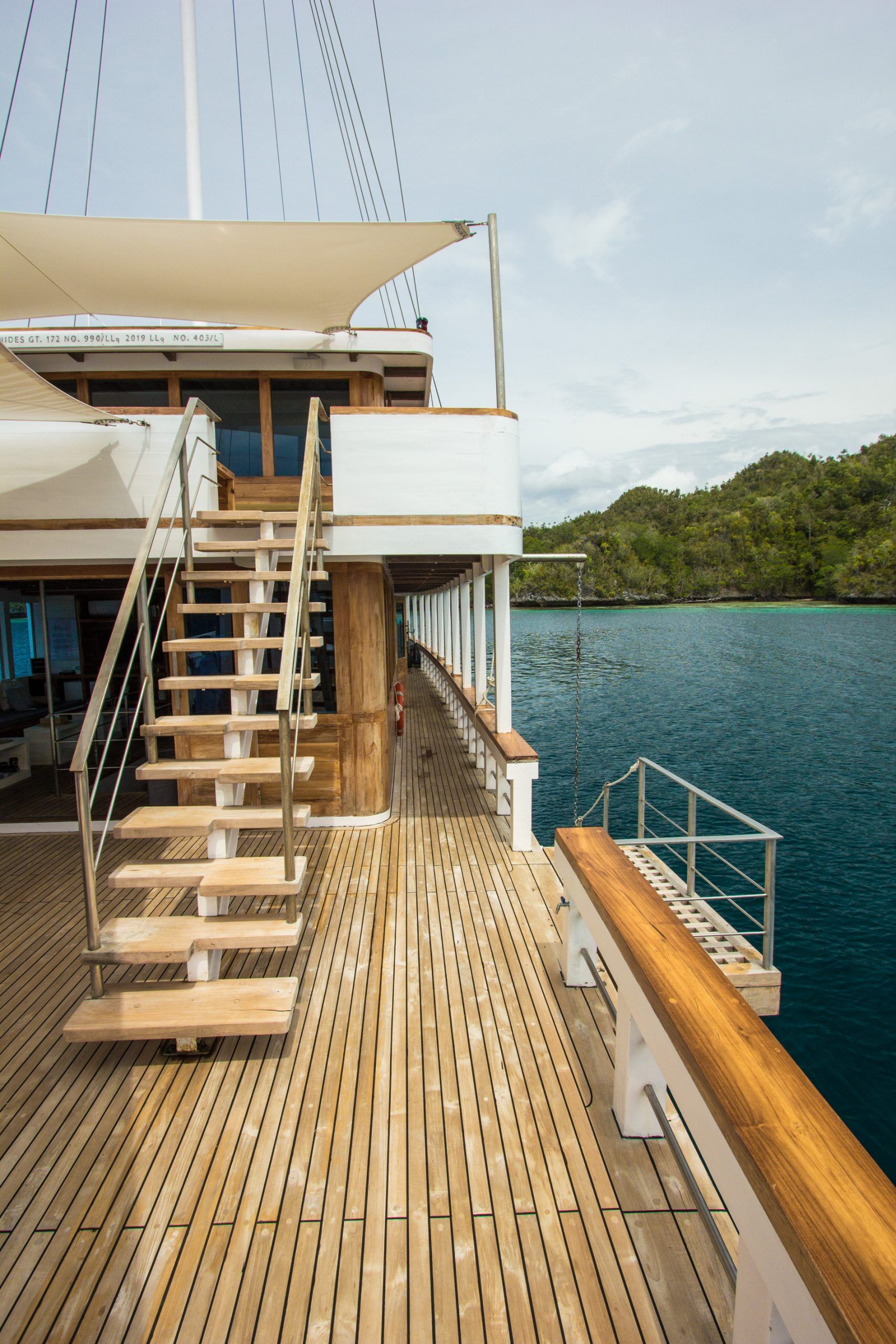 A wooden deck of a yacht with stairs leading to an upper level, railings along the edge, and a view of calm blue water and green hills in the background under a cloudy sky.