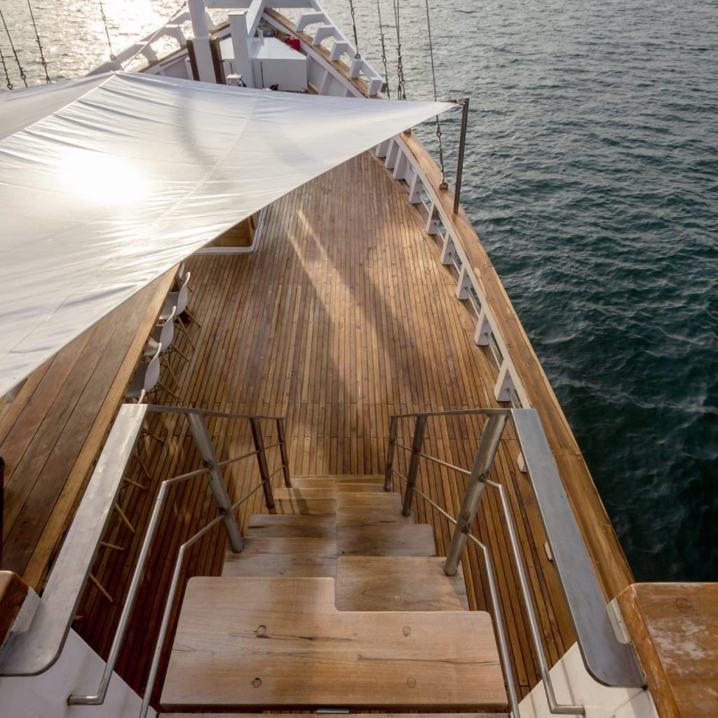 A view of a wooden yacht deck with a staircase leading down, a white canopy providing shade, and calm ocean water surrounding the boat. Sunlight reflects off the water and the deck.