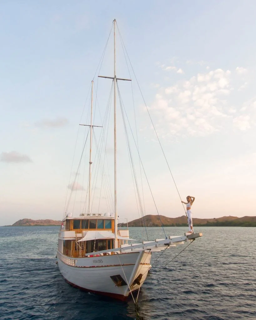 A person in white clothing stands at the bow of a large white sailboat on calm water, with hills and a partly cloudy sky in the background at sunset.