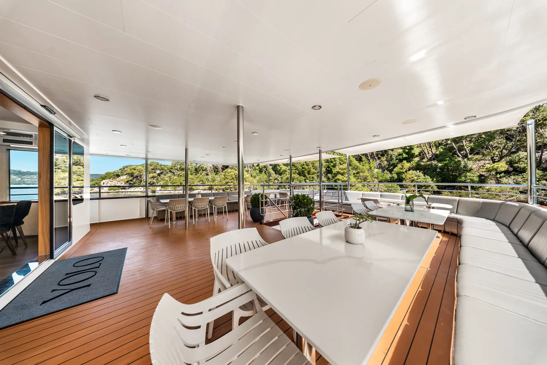 Spacious yacht deck with white tables, chairs, and a long cushioned bench. Surrounded by glass railings, it overlooks lush green hills and water, with natural light and open-air views.