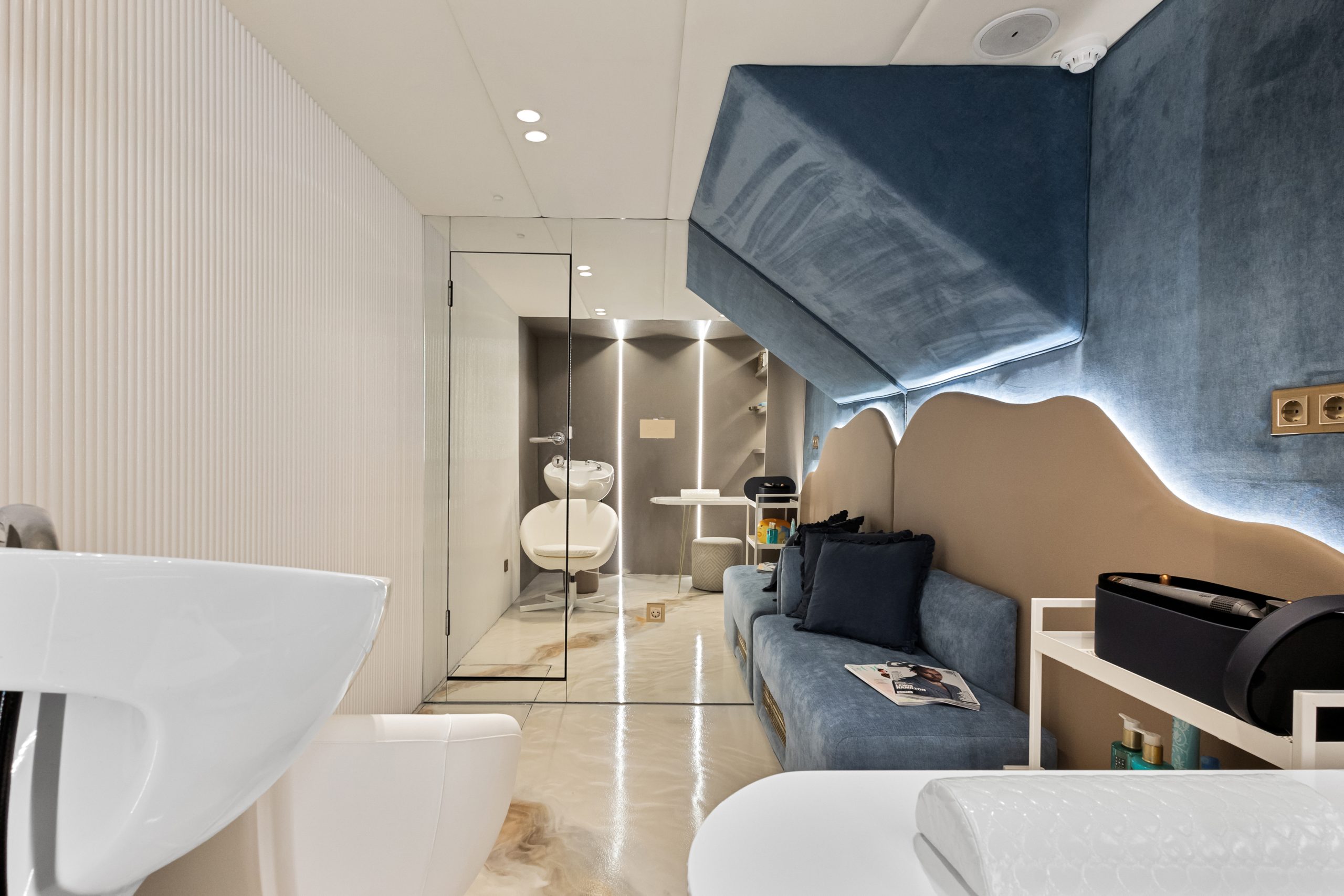 Modern salon interior with a glass-walled room containing a white salon chair and sink, blue velvet seating, decorative wall lighting, and shelves with beauty products and magazines.