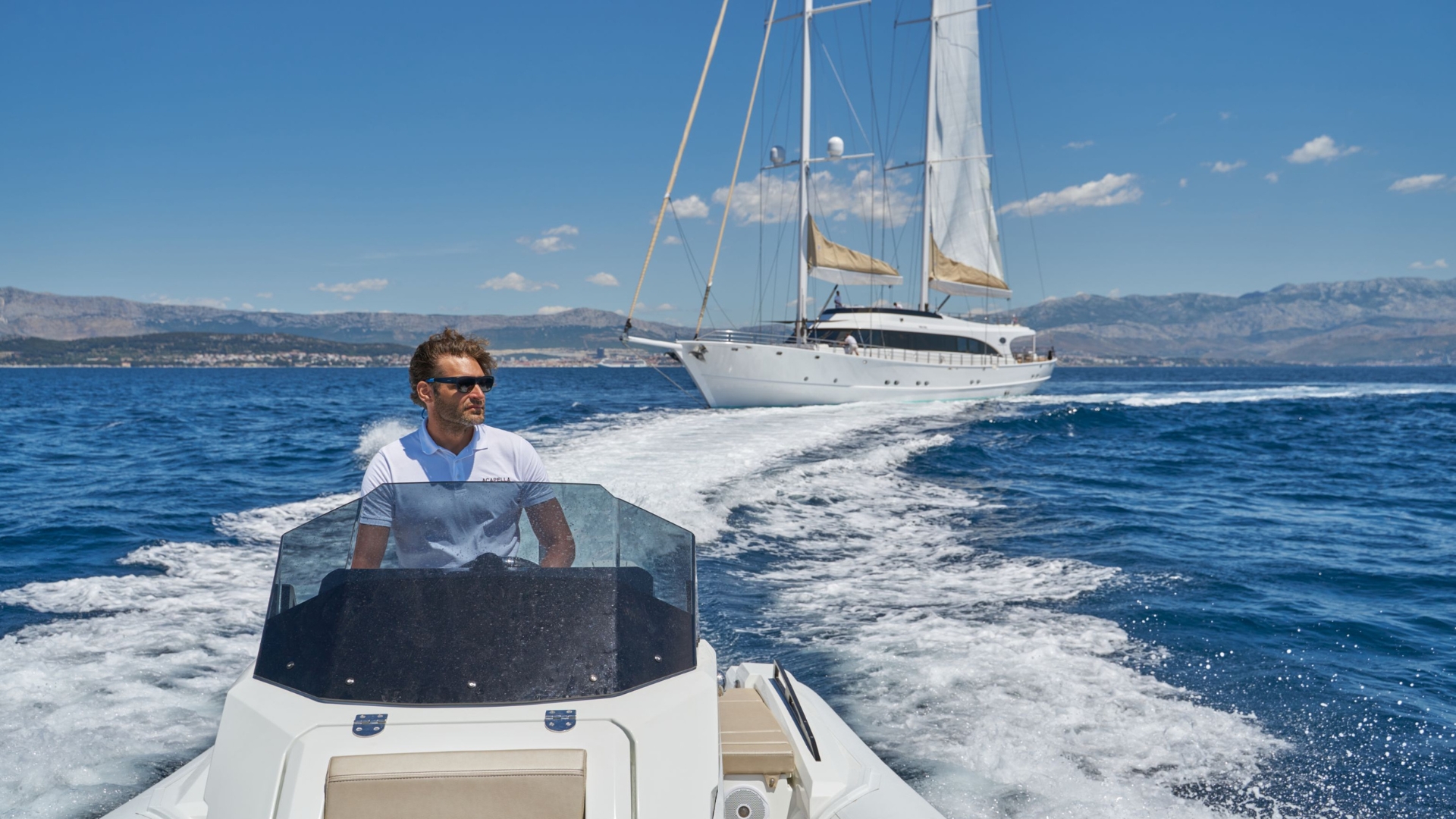 A man in sunglasses drives a small motorboat on blue water, with a large sailboat and distant mountains in the background under a clear, sunny sky.