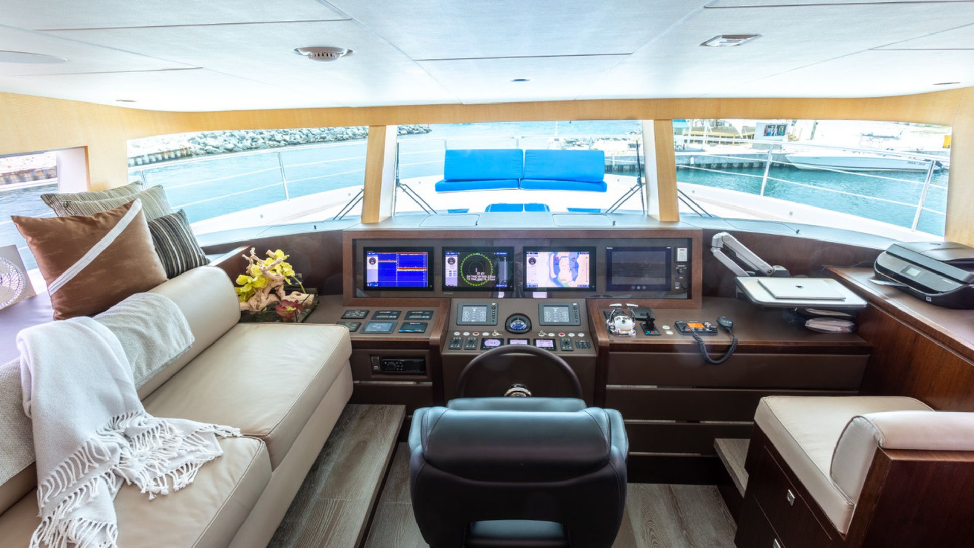 The image shows the interior of a luxury yacht cockpit with a modern control panel, comfortable cream-colored seating, decorative pillows, a throw blanket, and large windows overlooking the water and marina.