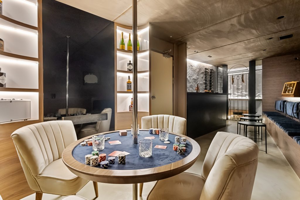 A modern, softly lit room with a round table set for poker, featuring cards, chips, and glasses. Plush white chairs surround the table, and shelves with bottles and a bar area are visible in the background.