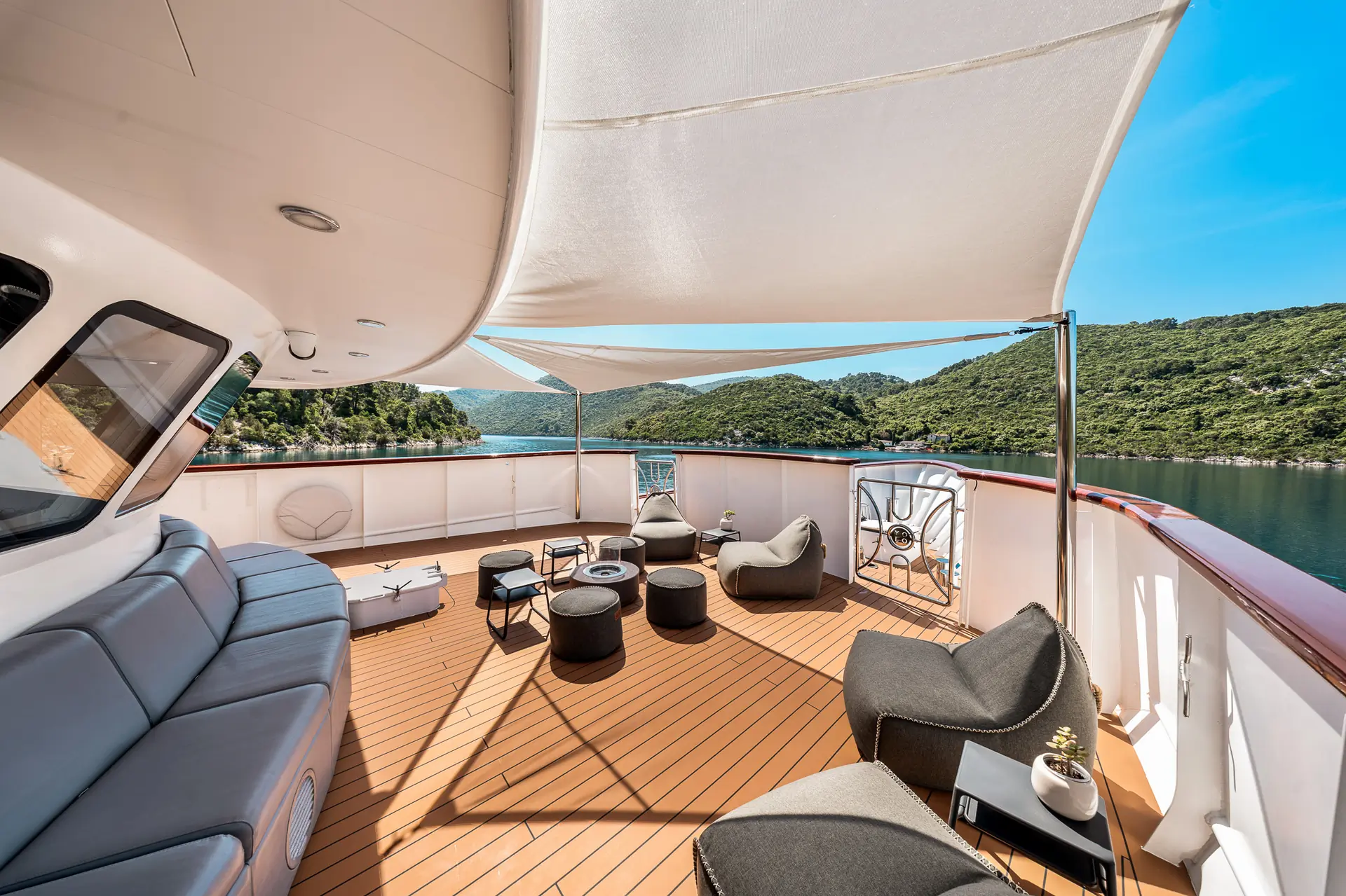Spacious yacht deck with modern lounge chairs, sofas, small tables, and a canopy overhead, overlooking calm water and lush green hills under a clear blue sky.