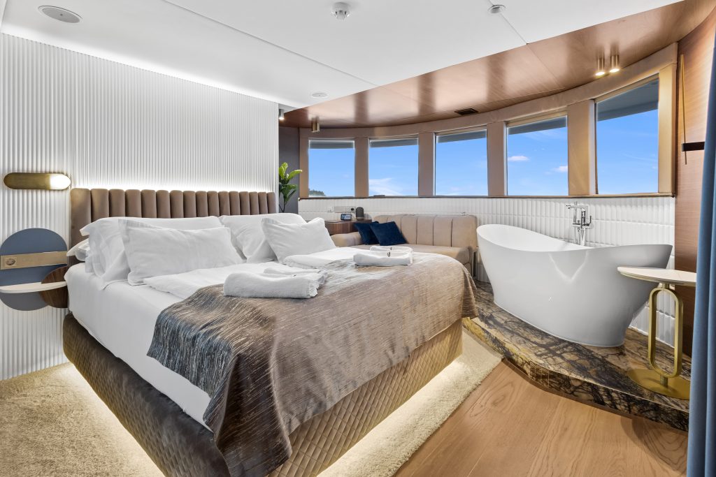 Modern hotel room with a large bed, folded towels, a freestanding bathtub, a beige sofa, and curved windows offering a scenic view. The room features warm lighting and stylish decor with wood and neutral tones.