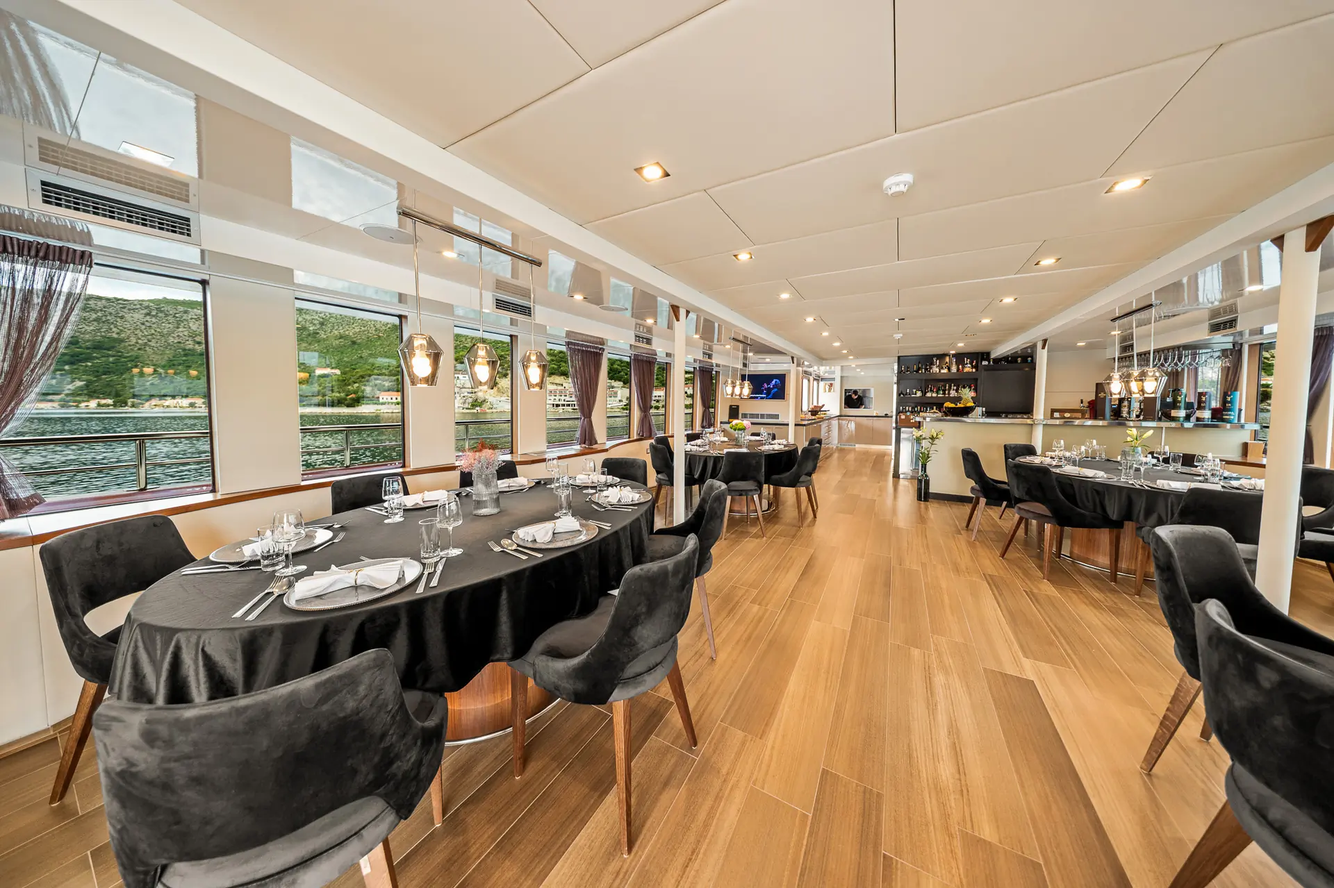 Modern dining area on a boat with round tables covered in black tablecloths, set with plates and glasses, black chairs, wooden floors, large windows overlooking water, and a bar in the background.