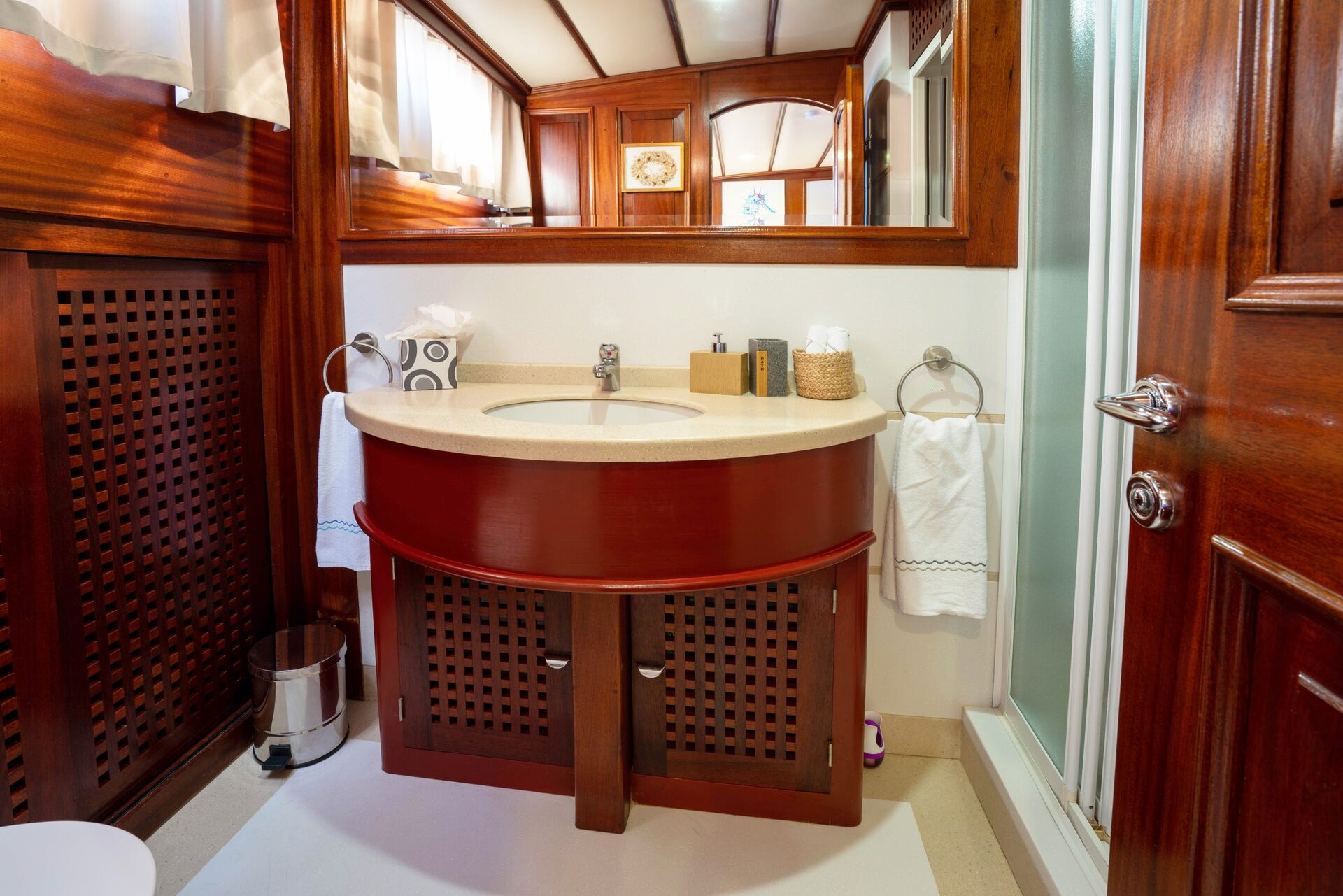 A bathroom with a wooden vanity, round sink, mirror, towel ring with white towel, tissues, toiletries, waste bin, and a glass shower door. The room features paneled wood walls and white accents.
