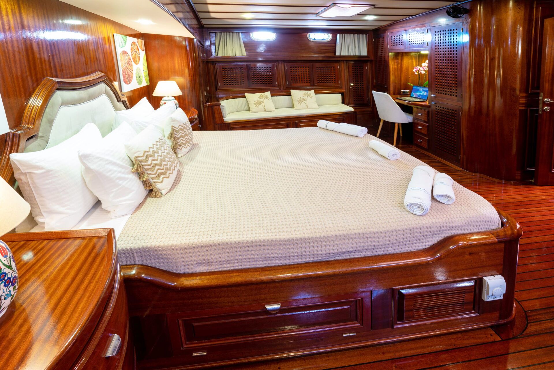 Luxurious yacht cabin with a large bed, wooden walls and furniture, soft lighting, a sofa, desk with chair, pillows, and neatly rolled towels on the bed; cozy and elegant atmosphere.