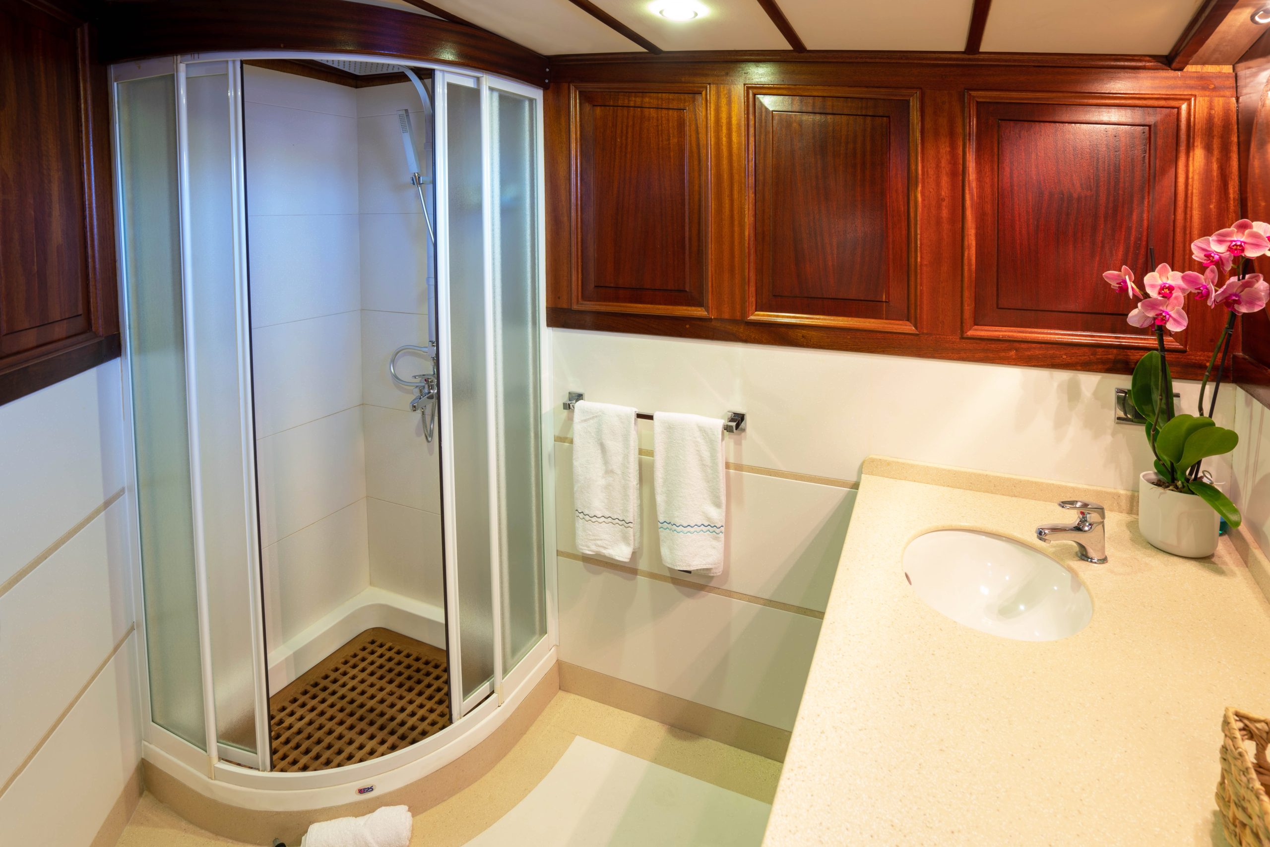 A bathroom with a corner shower cabin, white walls, wooden cabinets, a beige countertop with a sink, two white towels on a rack, and a pink orchid plant on the counter.