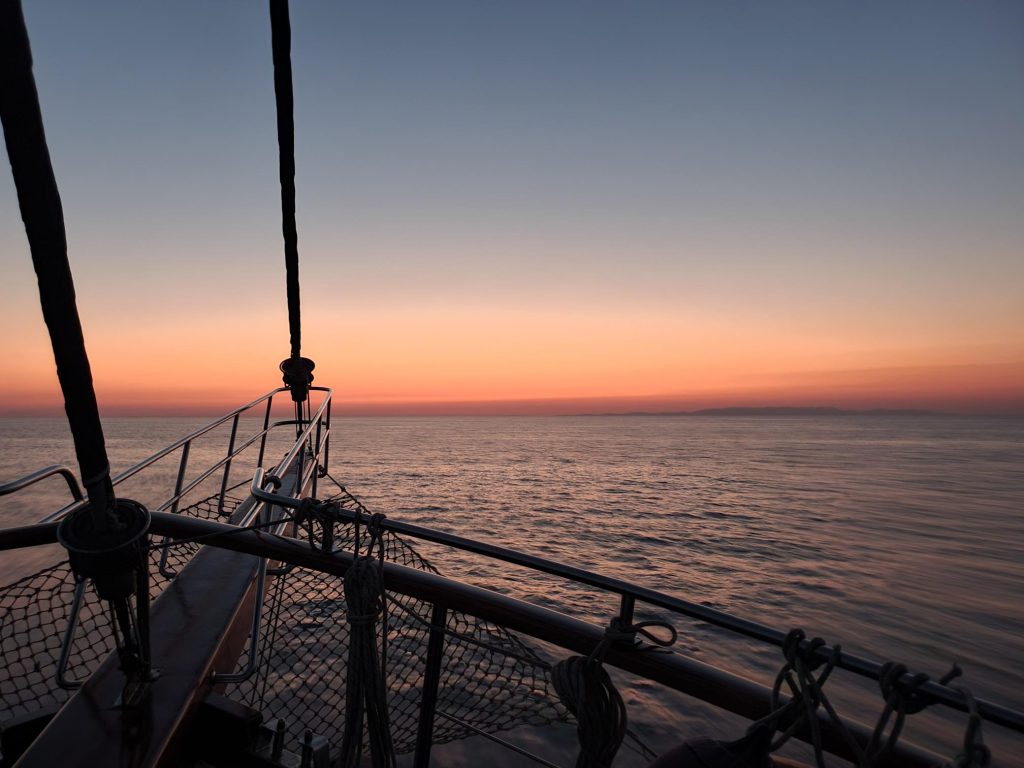 A boat sails on calm water at sunset, with the sky glowing in shades of orange and pink. The bow and railing of the boat are visible in the foreground, looking out over the tranquil sea.