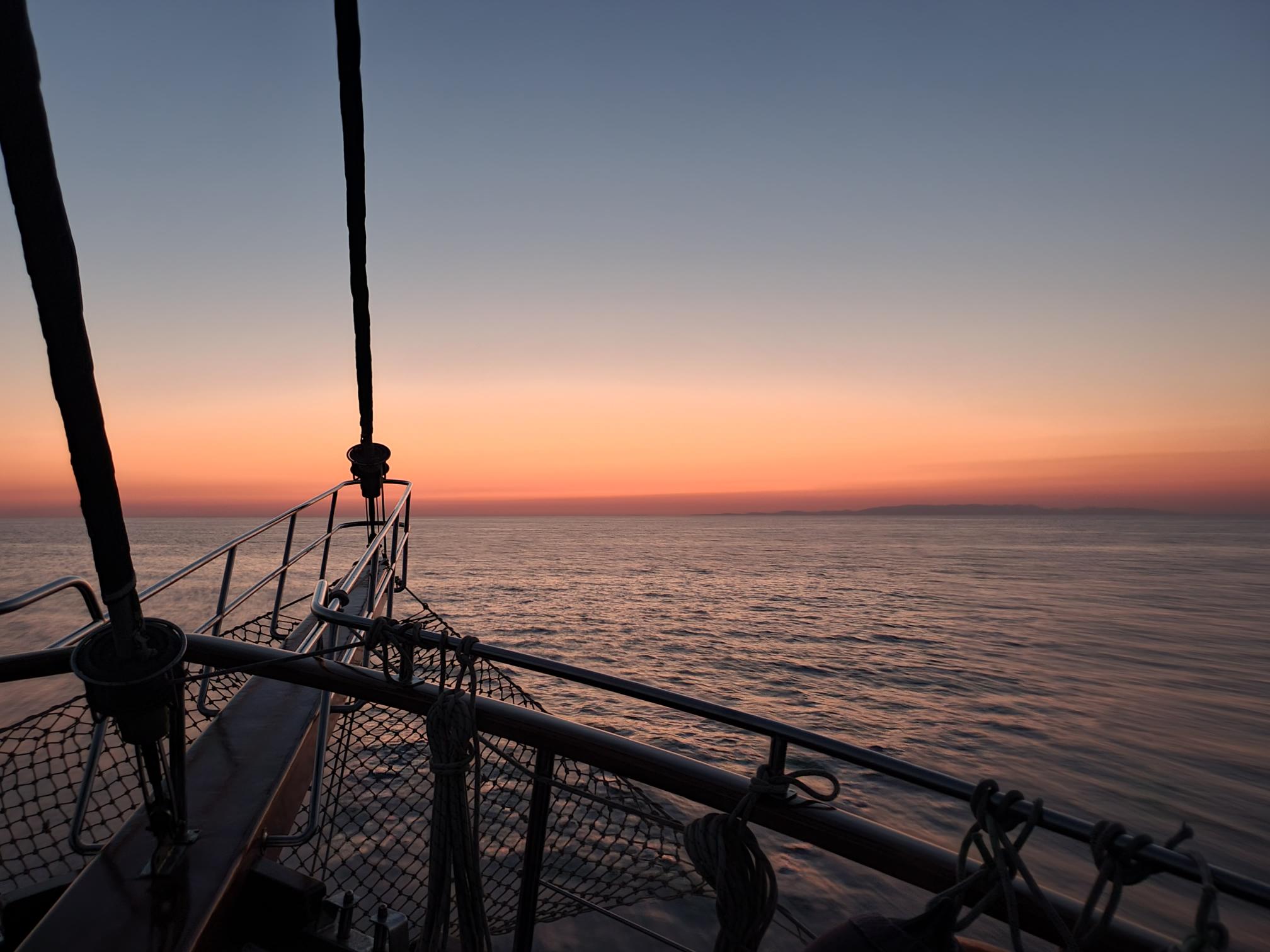 A boat sails on calm water at sunset, with the sky glowing in shades of orange and pink. The bow and railing of the boat are visible in the foreground, looking out over the tranquil sea.