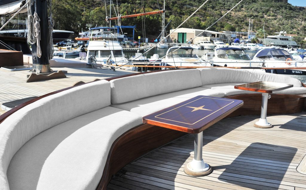 Curved cushioned seating and wooden tables on the deck of a yacht, anchored in a marina with several other boats and lush green hills in the background.